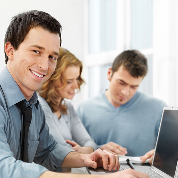 3 Junge Menschen sitzen vor einem Computer. Ein Mann schaut in die Kamera